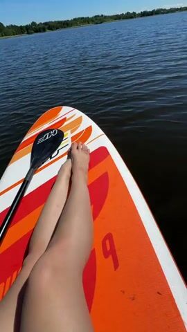 The video begins with a close-up, overhead shot of a person's bare legs resting on an orange and white paddleboard. The paddleboard is floating on dark, choppy water. The person's legs are light-skinned and appear to be crossed at the knees. A paddle is visible, lying on the paddleboard to the left of the person's legs. The water reflects the sunlight, creating shimmering highlights. In the background, a distant shoreline with trees and blue sky can be seen. The video pans slightly to the right, focusing on the water and the reflections on its surface, with the paddleboard remaining in the foreground. The overall impression is a peaceful outdoor scene, likely a lake or calm ocean on a sunny day, with the camera positioned on the paddleboard itself.