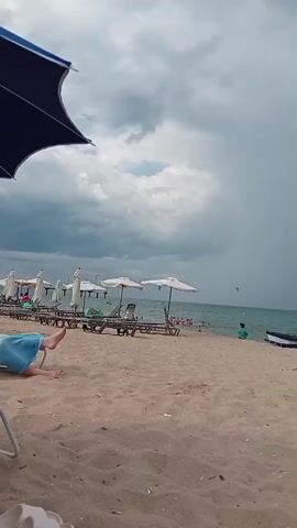 The video opens with a view of a sandy beach. A dark blue umbrella is visible in the foreground on the left. In the mid-ground, several white beach umbrellas are set up over lounge chairs. Some people are sitting on the chairs, and others are in the water. The sea is visible in the background, and the sky is overcast with dark clouds. A small boat is pulled up on the shore to the right. The overall atmosphere suggests a cloudy day at the beach, possibly with the threat of rain.