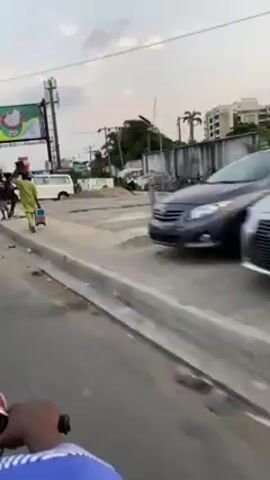 The video shows a street scene shot from the perspective of someone on a moving motorcycle. Initially, several people are seen walking on the sidewalk, some carrying items on their heads. As the motorcycle proceeds, the scene transitions to a roadside gathering. Numerous motorcycles are parked by the side of the road, and people are standing around. The area also has what appears to be merchandise laid out on mats. In the background are buildings, billboards, and some greenery. The sky is overcast.