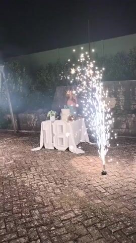 A close-up shot shows a table covered with a white tablecloth, adorned with a cake and the number 18 in large, white, illuminated block letters. In the foreground, a sparkler erupts with bright white sparks, illuminating the surrounding area. The background is blurred and dark, with hints of greenery and a stone wall. The scene appears to be an outdoor celebration at night, possibly a birthday party. The camera then pans up to show fireworks exploding in the dark night sky, with a few people visible in the lower portion of the frame, watching the display. The fireworks are colorful, with bursts of pink and white. The overall atmosphere is festive and celebratory.