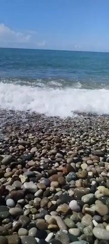This video shows a pebble beach with the ocean in the background. The waves are gently rolling onto the shore, creating a white foam that washes over the colorful pebbles. The water is a clear blue-green, and the sky is bright with a few scattered clouds. The sun appears to be high in the sky, suggesting it is daytime. The scene is peaceful and tranquil, evoking a sense of calm and relaxation. The pebbles are smooth and varied in color, ranging from light gray and white to darker shades of brown and black. Some of the pebbles appear wet from the waves, making their colors more vibrant. The overall impression is one of natural beauty and serenity.