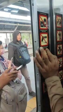 The video shows a view from inside a train car at a station platform. People are visible on the platform and some are boarding or disembarking. A hand is in the foreground, touching a set of vertical signs with red circles and diagonal lines, indicating prohibitions. These signs appear to depict items such as a lighter, a gun, and a knife, suggesting that these items are not allowed. A person wearing a hooded sweatshirt and a backpack is standing on the platform, facing the train. Another person wearing glasses and a mask is also on the platform, holding a phone and a backpack. The platform has yellow and blue markings. The overall scene depicts a public transportation environment with safety regulations in place.