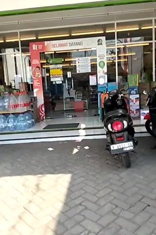 The video shows the exterior of a convenience store with its glass doors open. On the left, several large bottles of water are stacked on wooden planks. A black motorcycle is parked in front of the store. Inside the store, shelves are stocked with various products. A banner with the text "SELAMAT DATANG" (Welcome) is visible above the entrance. The floor in front of the store is paved with bricks.