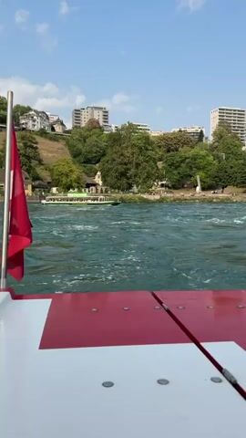 The video shows a boat moving on a wide, choppy green river. The camera is positioned at the front of a red and white boat, looking out over the water. In the distance, across the river, is a city built on a steep, green hillside. The city has many buildings, some tall and modern, others older and with red roofs. There are trees covering much of the hillside. Further to the right, a large waterfall cascades down rocks. On top of the hill, to the right of the waterfall, is a large, old castle or fortress with several buildings and towers. The sky is blue with scattered white clouds. The water is turbulent, with whitecaps and foam, suggesting a fast current. Another boat, green and white, is visible further upriver on the left. People are visible on the bank near the waterfall and castle.