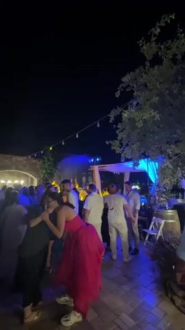 The video captures a nighttime outdoor gathering. The scene is illuminated by blue and white lights, with string lights visible overhead. A group of people, dressed in semi-formal attire, are seen mingling and dancing. Some individuals are wearing dresses, while others are in white shirts and pants. The setting appears to be a courtyard or patio with stone flooring and architecture in the background. The overall atmosphere suggests a festive event, possibly a party or celebration.