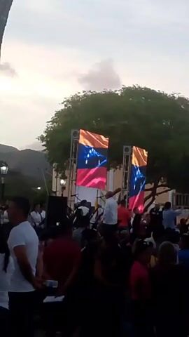 A crowd has gathered in a plaza, with two large video screens displaying the flag of Venezuela. Some people in the crowd are waving their arms in the air. There is a building behind the crowd with a sign that says "Museo" (museum in Spanish). The sky is a dusky color.