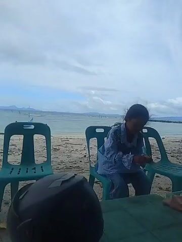 A young girl in a blue and white patterned shirt and blue pants sits on a teal plastic chair on a sandy beach. She is hunched over, looking down at her hands. Behind her, the ocean stretches to the horizon under a cloudy sky. To her left and right are two more teal plastic chairs. In the foreground, a green plastic table is visible, with a dark object, possibly a helmet, on its left side and a folded brown garment on its right. The girl briefly looks up at the camera before returning her gaze to her hands.