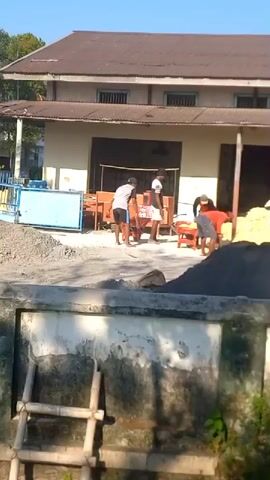 The video shows a group of men working outside a building. There are piles of gravel and what appears to be cement or sand. Two men are visible in the foreground, one wearing an orange shirt and the other in a white shirt. They are both interacting with what seems to be a wheelbarrow and possibly a mixing machine. Other figures are present in the background, suggesting a construction or materials handling site. The building in the background appears to be industrial or a storage facility with a corrugated metal roof and barred windows. The scene is outdoors during daylight, indicated by the shadows and bright lighting.