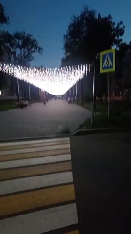 The video shows a pedestrian pathway at dusk, illuminated by strings of white lights hung overhead, creating a tunnel effect. The pathway is paved with interlocking stones. On the right side of the path, there is a pedestrian crossing sign. On the left side, there are trees and some benches. In the distance, several people can be seen walking along the pathway. The sky is a deep blue, indicating it is twilight. The camera is positioned near the beginning of the pathway, looking down its length.