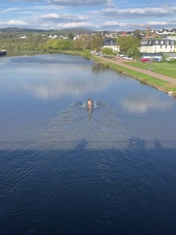 The video presents an elevated, wide shot of a four-person rowing shell with a coxswain, gracefully moving along a calm river under a bright, sunny sky. The boat is propelling away from the camera, leaving distinct V-shaped wakes on the dark blue water. Four rowers are visible, each with two oars, executing synchronized strokes, while the coxswain, wearing a yellow top, faces them from the stern.

The river is bordered by a lush, tree-lined bank on the left and a paved path on the right. Alongside the path are a mix of buildings with traditional European architecture, including white and pastel-colored houses, parked cars, and areas of green grass. In the background, a small town stretches along the river and up gentle, green hills. A large construction crane is visible in the distance on the right-hand side of the riverbank. The sky is a clear blue with scattered fluffy white clouds.

The camera's high vantage point, likely from a bridge, casts a large, dark shadow across the foreground of the river. The audio features the rhythmic sounds of oars splashing in the water, accompanied by clear voices giving instructions and counting in German, such as "Eins, zwei, drei, vier..." (One, two, three, four...). The scene depicts a focused rowing activity on a picturesque and tranquil day in Saarburg, Germany.