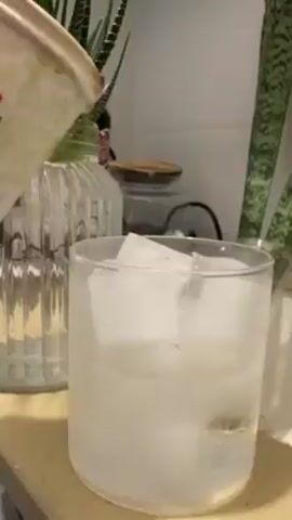 A person is pouring coffee from a paper cup into a clear glass filled with ice. The coffee is dark brown and forms a layer on top of the ice. There is a ribbed glass jar, a potted snake plant, and other kitchen items visible in the background. The surface is a light-colored countertop.