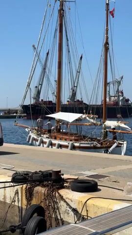 A schooner is docked at a pier. A person is visible on the deck of the schooner. In the background, a large cargo ship is moored with cranes extended. The sky is clear and blue. The pier is made of concrete and has various ropes, chains, and tires on it.