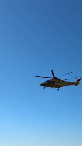 A yellow helicopter with red accents flies across a clear blue sky. The helicopter has a prominent tail rotor and main rotor blades, which are blurred from rotation. Its landing gear is extended. The scene is captured from a low angle looking upwards.
