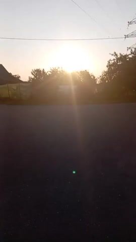 The video shows a rural road scene at sunset. The sun is low in the sky, casting a warm glow and creating lens flare. A power line tower and poles are visible against the sky. Trees line the road, and a small house is situated behind the trees. The road itself appears to be asphalt with some damage, and a small puddle of water is visible on the side. The video pans slowly to the right, showing more of the road and the surrounding vegetation.