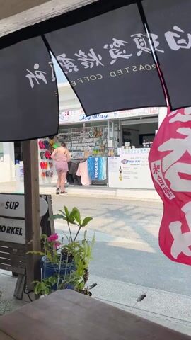A woman in a pink top and shorts is seen walking towards a beach shop called "My Way". The shop is decorated with colorful flip-flops and souvenirs. A banner advertising "Roasted Coffee" hangs overhead. The scene is viewed from a shaded area, with a wooden table and potted plant in the foreground. A red flag with Japanese characters is partially visible on the right. The environment appears to be a sunny coastal area.