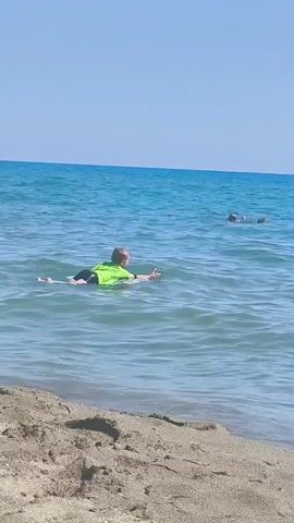This media file captures a sunny day at the beach, likely in Castiglione della Pescaia, Italy, given the location context. The scene is set in the calm, blue waters of the sea, with a sandy shoreline in the foreground. The sky is a clear, bright blue, indicating good weather and likely midday.

The primary subjects are two individuals in the water, both appearing to be children, engaged in recreational activities. The child closer to the shore is lying on a boogie board, wearing a bright green rash guard. This child is positioned to face the open sea, suggesting they might be waiting for a wave or observing others. Further out in the water, another child is visible, also appearing to be in the water, possibly with a flotation device. The presence of boogie boards indicates a day of fun and water sports.

The activity is leisure and play in the ocean. The gentle waves lapping at the shore and the overall peaceful atmosphere suggest a relaxed and enjoyable experience. The clear water and bright sunlight contribute to a feeling of summer vacation and outdoor enjoyment. There are no visible text elements in this media file.