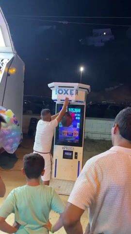 A man in a white t-shirt and white shorts is playing a boxing arcade game. He is standing to the left of the game and has just finished punching the hanging bag. The game machine has a digital display showing scores. A young boy in a green t-shirt is standing in front of the game, watching the man. Another person, mostly obscured from view, is standing to the right of the game, also watching. The scene appears to be outdoors at night, with some lights and the dark shapes of vehicles in the background.