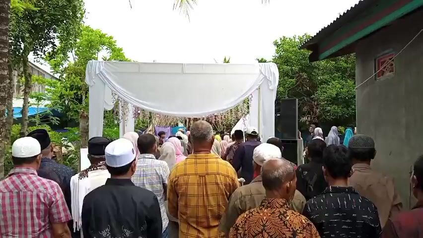 A group of people, mostly men wearing hats and some women in headscarves, are gathered outdoors, facing a white draped archway. The archway is decorated with hanging flowers. Behind the archway, more people are visible, suggesting an event or ceremony is taking place. The setting appears to be a rural or residential area, with trees, foliage, and simple buildings in the background. A speaker is positioned to the right of the archway. The overall atmosphere seems to be one of attendance or observation.