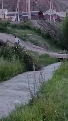 The video shows a person on a dirt path overlooking a fast-flowing, murky river. The person, dressed in white, is running up the path away from the river. The surrounding area is hilly and covered with green grass and bushes. In the background, there are houses and utility poles, suggesting a rural or semi-urban setting. The sky appears overcast, and the light suggests it could be dusk or dawn. The overall mood is serene and natural, with the rushing water creating a sense of movement and life. The presence of houses and utility poles indicates human habitation in the area. There is no visible text in the video. The video appears to be filmed in Naryn City, Kyrgyzstan.