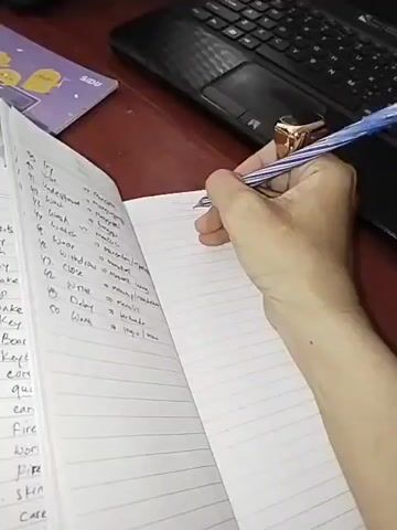 The video shows a close-up of a person's hand holding a blue pen and writing in a notebook. The notebook is open, and on the left page, there is a list of numbered words followed by their Indonesian translations. The person's right hand is writing on the right page, which is blank lined paper. The hand is wearing a large gold ring with a dark stone. In the background, a laptop keyboard and a purple object with yellow graphics are visible. The lighting suggests an indoor setting.