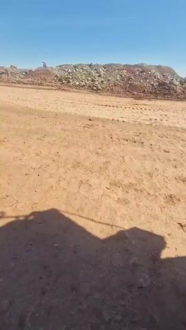 A very large yellow dump truck is driving on a dirt road. The truck is heavily loaded with brown rocks or dirt. The sky is clear and blue. There is a pile of brown and green rocks in the background. The sun is shining, casting a shadow of the dump truck on the road.