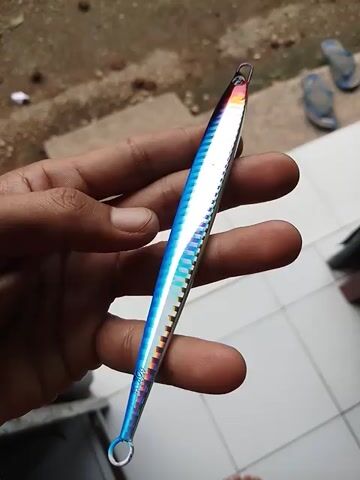 Here is a concise description of the video:

The video shows a hand holding a fishing lure. The lure is long and slender, with a silver base and colorful stripes in blue, green, and yellow. It has an eyelet at each end for attaching fishing line and hooks. The hand rotates the lure to show different angles, with a tiled floor and the person's feet visible in the background.