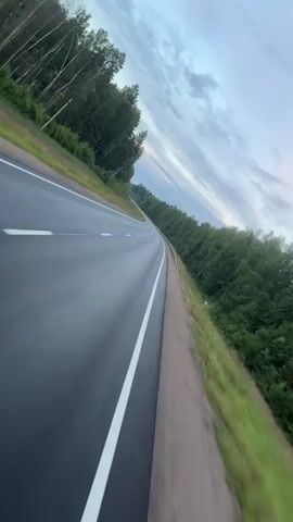 A POV shot from a moving vehicle shows a winding asphalt road through a forest. The road has a white dividing line and dashed white markings. The sky is cloudy. The vehicle appears to be on the right side of the road.