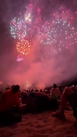 The video captures a fireworks display viewed from a sandy beach at night. Silhouettes of people are visible in the foreground, sitting or lying down on the sand, facing the sky. The sky is filled with a variety of colorful fireworks, predominantly red, gold, white, and purple, exploding and leaving trails of smoke. The light from the fireworks illuminates the smoke and casts a reddish glow on the scene. The overall atmosphere suggests a celebratory event.