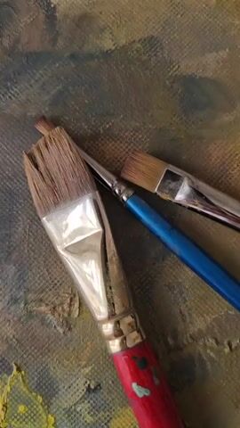 Three paint brushes are lying on a textured surface. The largest brush in the foreground has a wide, flat head with natural bristles and a red handle. To the right and behind it, a smaller brush with a blue handle and natural bristles is visible. A third, very thin brush with natural bristles and a metallic ferrule is positioned behind the other two. The surface beneath the brushes appears to be a canvas with a textured, mottled painting in shades of green, brown, and yellow. The lighting is soft, and the camera is positioned directly above the brushes, looking down.