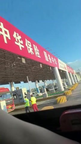 The video shows a view from inside a vehicle at a toll plaza. A large red and white banner with Chinese characters is visible overhead. The banner has the text "中華保險 服務中華" which translates to "China Insurance Service China". Below this, there are signs with numbers and logos. The toll plaza has multiple lanes, with some vehicles, including trucks and cars, passing through. Yellow and black striped barriers are present in the lanes. The sky is blue with some clouds, suggesting daytime. The interior of the vehicle is partially visible in the foreground, including a dark-colored dashboard and a red object with USB ports.
