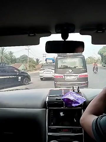 A first-person view from inside a vehicle shows a traffic jam on a road. The dashboard of the car is visible in the foreground. Through the windshield, several vehicles are seen on the road, including a black SUV, a white SUV, and a black minivan. A motorcycle rider is visible in the distance to the right. The environment appears to be outdoors with trees and utility poles visible. The sky is overcast. The video is about 5 seconds long.