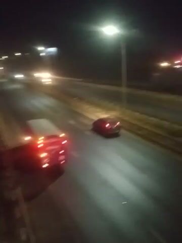 The video shows an aerial view of a highway at night. There are several vehicles on the highway, including what appears to be a semi-truck with its brake lights on and several other cars. The scene appears to be a traffic incident, as there are emergency lights flashing from some of the vehicles. The road is wet, suggesting it may have been raining. The video is shaky and blurry, indicating it was likely recorded from a moving vehicle or drone.