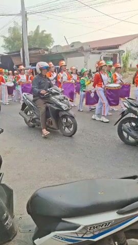 A street parade is taking place. A group of young people in matching orange, white, and green uniforms are marching and playing drums. Several motorcycles are also present, some carrying riders and passengers, and one is in the foreground. The street appears to be paved, with buildings and some greenery visible on the sides. The sky is overcast. The overall atmosphere suggests a community event or celebration.