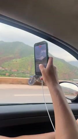 A close-up shot from inside a vehicle shows a person's hand holding a smartphone, which is actively recording a video. The phone, encased in dark green, displays a camera interface with a red recording timer that progresses from 00:00:01 to 00:00:05, and zoom controls (0.5x, 1x, 2.5x). A white charging cable is connected to the phone.

Through the vehicle's window, a mountainous landscape unfolds, featuring green and reddish-brown slopes under a cloudy sky. A road or highway with a guardrail is visible between the vehicle and the mountains. The setting appears to be along a scenic route in Taljarft, Morocco. Background music is audible throughout the video.