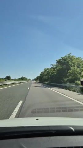 The video shows a first-person perspective of driving on a highway on a clear, sunny day. The road is asphalt with white lane markings. On the left side of the road, there is a metal guardrail and then fields with some trees. On the right side of the road, there are trees and bushes. In the distance, there are other vehicles on the highway and a billboard that says "mobile.bg". The sky is blue with some wispy clouds. The dashboard of the vehicle is visible at the bottom of the frame.