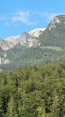 A time-lapse video shows a mountainous landscape with a clear blue sky and scattered clouds. The foreground features a dense forest of green trees. Behind the forest, the midground showcases the lower slopes of mountains covered in a mix of trees and rocky outcrops. The background displays the majestic peaks of higher mountains, some of which are snow-capped, with rugged, rocky faces. The sun shines brightly, casting light and shadows across the terrain, suggesting a clear day. The video captures the subtle changes in light and shadow as time progresses, emphasizing the scale and natural beauty of the mountain environment.