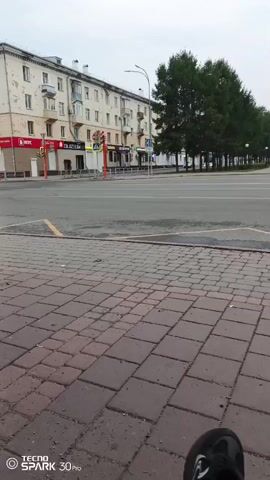 The video opens on a street with a multi-story building on the left and a line of trees on the right. In the foreground, there is a paved sidewalk made of red and gray square bricks. A person's legs and feet, wearing dark pants and black shoes with white accents, are visible in the bottom right corner of the frame, suggesting the camera is held from a seated or low vantage point. The street is wide and appears to be an intersection with crosswalks and traffic signals. Cars and a bus stop shelter are visible in the distance. The sky is overcast, indicating potentially cloudy or early morning weather. The scene is quiet and there are no people readily apparent in the immediate vicinity of the camera. The video then pans slightly to the right.