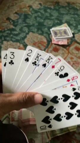 A hand holds a fan of playing cards, with a 10 of spades prominent in the foreground. Behind the fan, other cards are visible, including a 9 of clubs and a King of spades. In the background, a patterned green rug is visible, with stacks of paper money and playing cards scattered on it. The lighting suggests an indoor setting.