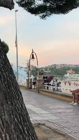 A group of people are gathered near a beach promenade. Fireworks are being set off in the background, creating plumes of white smoke. In the foreground, a man sits on a bench, appearing to observe the fireworks. Several other people are seated or standing nearby. The scene takes place at dusk, with a pastel-colored sky. A lamppost with an ornate design stands in the middle ground. Trees and buildings are visible in the background.