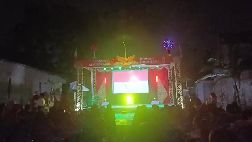 A nighttime outdoor stage setup is visible, featuring a large screen displaying the Indonesian flag. The stage is adorned with red and white decorations, including curtains and flags, and illuminated by stage lights and decorative lights on top of the scaffolding. A golden eagle emblem is prominently displayed above the screen. A crowd of people, mostly silhouetted, is seated in the foreground, facing the stage. The background appears to be a dark, open area with some structures and trees, suggesting a public gathering or event. The overall atmosphere is festive and celebratory.