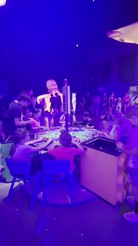 A group of children are gathered around a large circular table covered in colorful Lego bricks, building something together. The table is lit from above with purple lights, and there are larger Lego creations and figures visible in the background, including what appears to be a giant octopus statue. Adults are also present, supervising the children and participating in the activity. The environment is dimly lit, with a focus on the bright colors of the Lego bricks.
