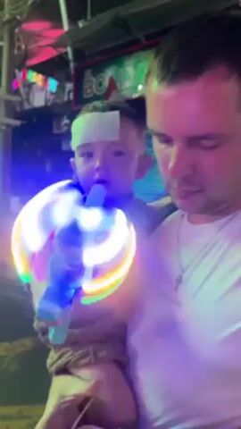 Here is a description of the video:
A man with short hair and a white t-shirt is holding a small child in his arms. The child, who has a bandage on their forehead, is looking at a spinning, light-up toy that the man is holding. The man has a backpack on and a tattoo on his forearm. The background shows a street scene at night, with signs and lights, including a sign that says "Bollywood" with an Indian flag on it. The man appears to be looking at the toy as well.