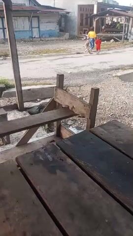 The video opens with a close-up view of weathered, dark wooden planks that appear to be part of a table or seating area. Beyond these planks, the scene transitions to an outdoor environment with a paved road and several low-rise buildings. In the background, two children, one wearing a yellow shirt and the other in a red shirt, are walking away from the camera. A person, possibly an adult, is seen walking behind them. Further down the road, a motorcycle with a sidecar is approaching, carrying at least one passenger. The buildings in the background are simple structures, some with visible paintwork. The ground is uneven and appears to be covered in gravel or dirt in places. The overall impression is of a rural or semi-urban setting.