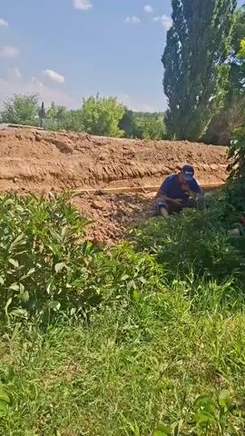 The video shows a group of four men outdoors on a sunny day. In the foreground, there is lush green grass and foliage. In the middle ground, a dirt trench is visible, with what appears to be a wooden frame or structure along its edge. Three of the men are seated or kneeling near the trench, surrounded by vegetation. One man, wearing a blue shirt and a baseball cap, is actively working with his hands in the trench. Another man, wearing a yellow safety vest over a blue shirt and a baseball cap, is also seated near the trench. A third man, wearing a yellow safety vest and dark pants, is standing and looking down at something in his hands, possibly a phone. A fourth man, wearing a red shirt and a yellow safety vest, is lying down or resting near the white structure on the right. The background features a dirt embankment and trees. The overall scene suggests manual labor or construction work is taking place in a rural or semi-rural environment. The presence of safety vests indicates a work setting. The time of day appears to be daytime, with clear skies and bright sunlight. Visible text on the yellow vest of the standing man reads "NOBUCONST".