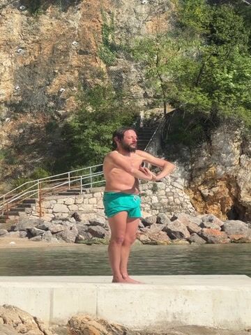 The video shows a man standing barefoot on a concrete platform by the water. He is wearing green swimming trunks and is shirtless. He is performing arm exercises, bringing his hands towards his shoulders. Behind him is a rocky cliff face with vegetation and a set of stone steps with a white railing leading up the cliff. The water is calm, and there are rocks along the shoreline. The sky is bright.