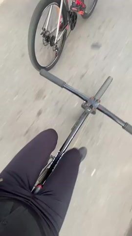 The video captures a first-person view of someone cycling on a paved road. The rider's legs, dressed in black leggings, are visible, along with the black handlebars and frame of their bicycle, which features the brand name "VENZO" in white text. Another cyclist is seen ahead and to the left, with their rear wheel, white-spoked wheels, and shoes with red accents visible. The two individuals are actively cycling. The presence of distinct shadows and warm sunlight indicates that it is either morning or late afternoon/evening, under clear, sunny weather conditions. The scene takes place outdoors on what appears to be an asphalt or concrete surface. The location context for this activity is Monte Grande, Argentina.