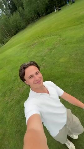 A man in a white shirt walks while holding a golf bag and cart. He walks on a golf course with green grass and trees. He stops and pulls the cart. The cart is full of golf clubs and a few hats. The background of the golf course has green hills and a cloudy sky.