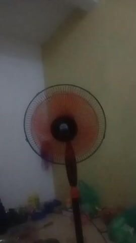 A stationary oscillating fan is shown from the chest up. The fan blades are a translucent orange and appear to be spinning. The fan has a black grille and a black circular motor housing. The fan is positioned in front of a light yellow wall. The lower portion of the fan's stand is visible, which is black with an orange band. In the foreground, at the bottom of the frame, is a blurry collection of various objects, including what appear to be plastic bags, a metal bowl or container, and other assorted items. The lighting is dim and appears to be artificial, possibly from an overhead source. The video quality is low, and the image is somewhat grainy and out of focus, particularly in the foreground.