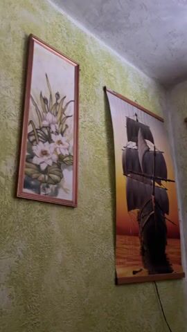 The video shows a corner of a room with three framed artworks on a textured light green wall. On the left is a tall, narrow framed painting of white lilies with green leaves. To its right, hanging vertically, is a large tapestry-style artwork depicting a dark sailing ship with three masts against a vibrant orange and yellow sunset over the water. Further to the right, another similar tapestry artwork is hung, showing a narrow street lined with buildings and a lamppost, with a body of water and mountains in the background. The ceiling is visible at the top and appears to be textured as well, with a slight off-white color. Two power cords are visible hanging from the bottom edges of the two tapestry artworks.