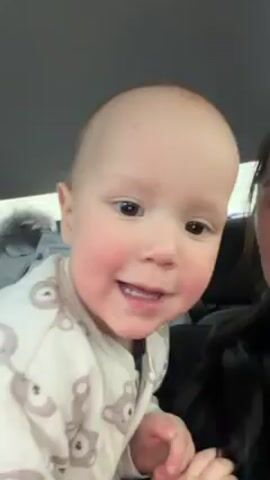 A baby is being filmed in a car. The baby is wearing pajamas with bear prints. The baby smiles and makes a "raspberry" sound. A person in a black shirt is sitting next to the baby. A jacket can be seen in the background. The interior of the car is visible.